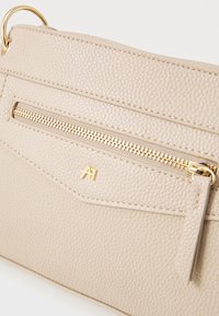 Beige tekstureret lædertaske med en guld lynlås, med en frontlomme og en subtil logo detaljer. Glat overflade og minimalistisk design.