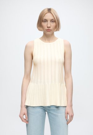 Vrouw met kort blond haar die een mouwloze crèmekleurige gebreide top en lichtblauwe jeans draagt, staand tegen een effen lichte achtergrond.