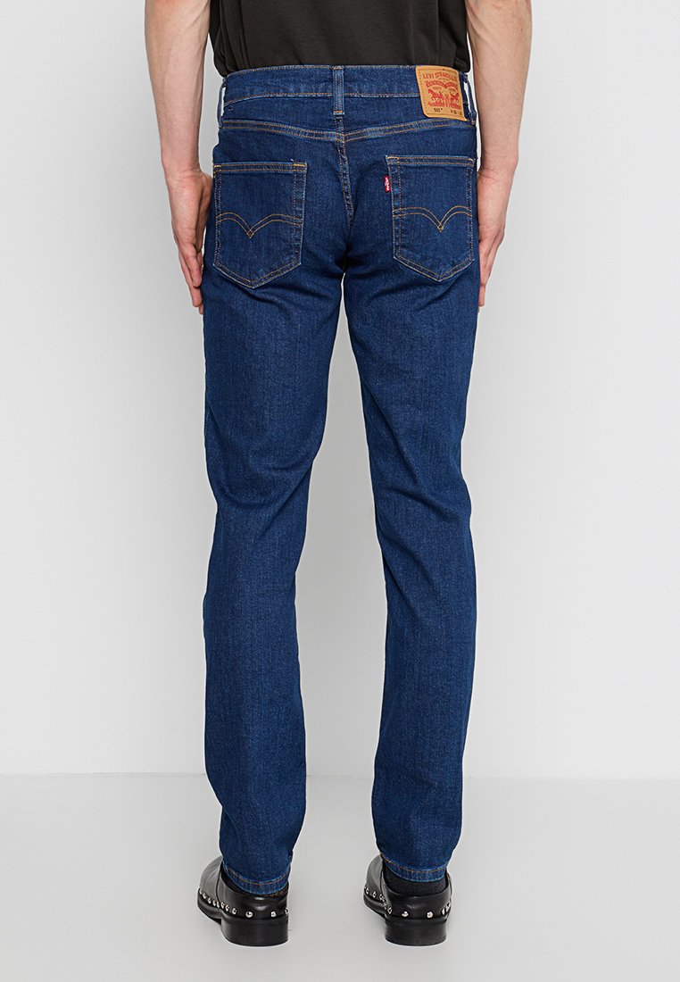 Man wearing dark blue Levi's jeans, standing with arms relaxed, visible leather brand patch and back pockets with stitching detail.