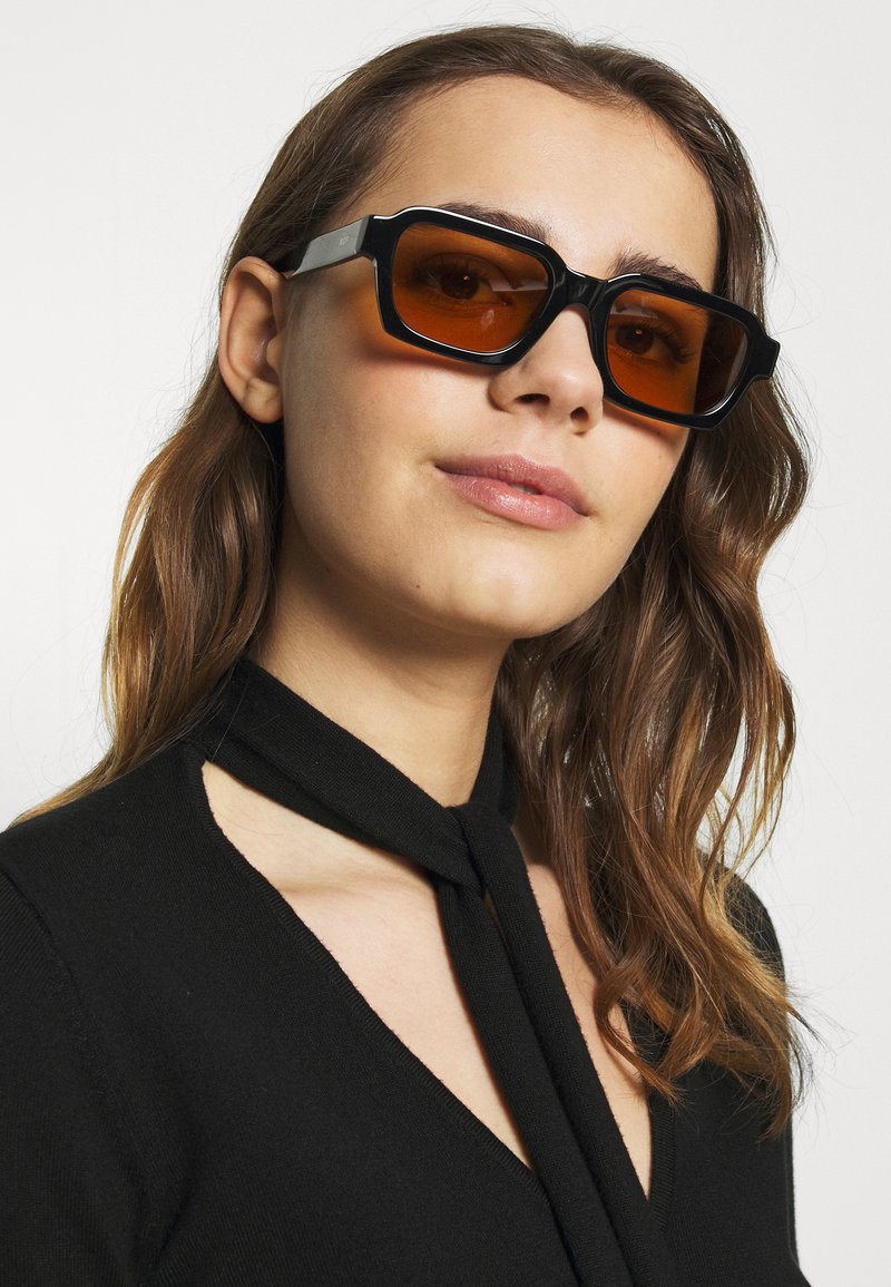 Occhiali da sole neri rettangolari con montatura spessa e lenti color ambra. Il modello indossa una maglietta nera con un dettaglio a fiocco sullo scollo.