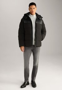 Giacca a puffo nera con cappuccio, chiusura con zip e design trapuntato. Indossata sopra un maglione grigio chiaro a trama e jeans grigi, abbinata a stivali neri.