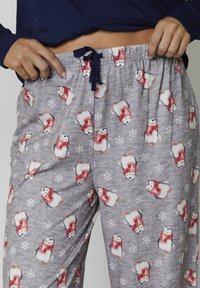 Persona che aggiusta pantaloni del pigiama grigi con motivi di pinguino con sciarpa rossa e fiocchi di neve bianchi, indossando una maglietta a maniche lunghe blu navy.