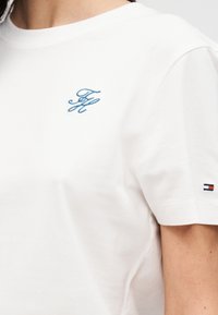 T-shirt en coton blanc à manches courtes ; avec un logo brodé bleu sur la poitrine et un petit emblème multicolore sur la manche.