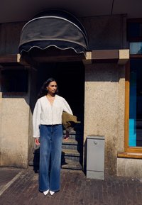 Valge tekstuuriga lühike kampsun, kõrge vöökohaga helerohelised kellukad teksapüksid ja valged teravnurksed kontsad. Modell hoiab õlalehte rohelist mantlit. Väljas keskkonnas.