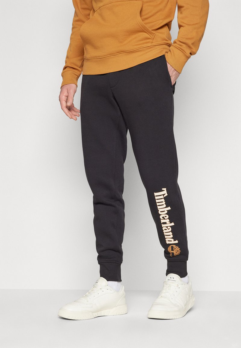 Mörkgrå träningsoverallsbyxor med en tapered passform, med en stor "Timberland"-logotyp i beige på nederbenet. Tillverkade av mjukt tyg med ribbade muddar.