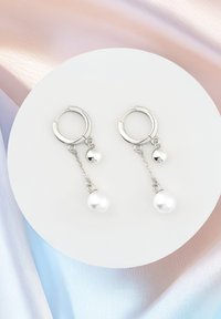 Silverörhängen med gångjärn prydda med en kedja som har en sfärisk pärla och ett hängande vitt pärlvedhänge, mot en mjuk, mångfärgad bakgrund.