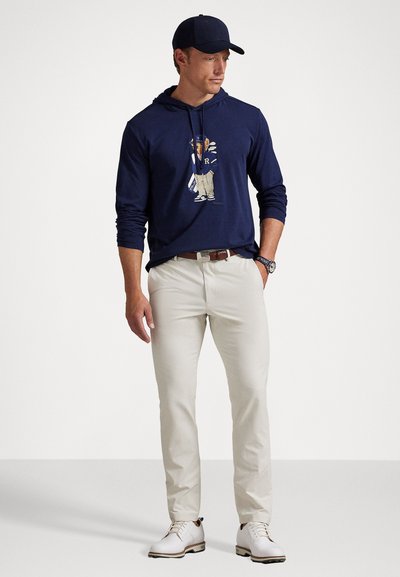 Man in een marineblauwe baseballcap en een hoodie met een cartoonfiguur, beige broek, een bruine riem, witte schoenen en een horloge, staande tegen een witte achtergrond.