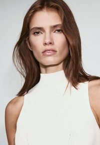 Top alb, fără mâneci, cu gâtul înalt, având o textură cu nervuri. Modelul are părul lung, brunet și un machiaj neutru.