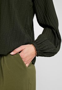 Blusa verde scuro con texture e maniche ampie, pantaloni verde oliva con dettaglio a tasca, accompagnati da un sottile braccialetto dorato al polso.