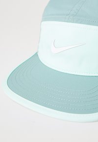 Lyseblå og mintgrønn Nike-cap med hvit swoosh-logo på frontpanelet, detaljert søm på den buede skjermen, satt mot en enkel bakgrunn.