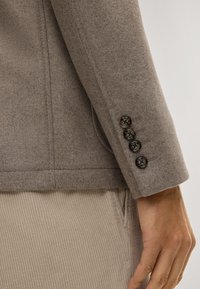 Hellbrauner Wollblazerärmel mit vier schwarzen Knöpfen, der eine glatte Textur und ein maßgeschneidertes Design hervorhebt. Der Hintergrund zeigt beige Cordhosen.