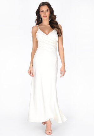 Nő, aki hosszú, fehér ujjatlan ruhát visel vékony pántokkal és gyöngy fülbevalókat, magabiztosan sétál fehér háttér előtt.