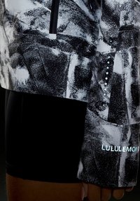 Haut de sport à manches longues à motif noir et blanc avec fermeture éclair frontale, passages pour les pouces, points réfléchissants et texte "Lululemon" sur la manche.