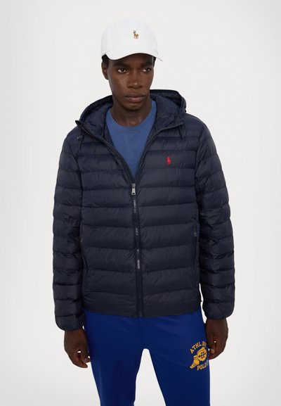 Uomo con cappello bianco, giacca a vento blu navy con logo rosso, t-shirt blu e pantaloni sportivi blu con design atletico giallo stampato.