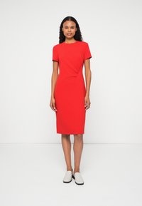 Piros, testhezálló ruha rövid ujjakkal, kerek dekoltázzsal, oldalsó redőzéssel és térd alá érő szoknyával. A modellen szürke slip-on cipők vannak.