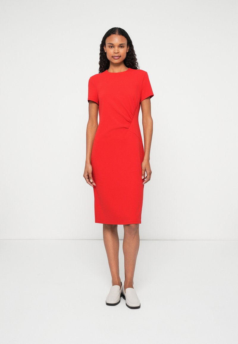 Piros, testhezálló ruha rövid ujjakkal, kerek dekoltázzsal, oldalsó redőzéssel és térd alá érő szoknyával. A modellen szürke slip-on cipők vannak.