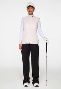 Ljusrosa quiltad väst med hög krage, bärs över en vit långärmad skjorta. Har svarta golfbyxor och vita sneakers, håller i en golfklubba.