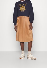 Felpa blu navy con logo dorato, gonna midi beige plissettata e sneakers beige. Tessuto morbido, vestibilità rilassata e dettaglio testurizzato della gonna.