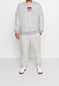 Felpa grigia con logo "GANT" e design con scudo, abbinata a pantaloni della tuta grigi coordinati e sneakers bianche con dettagli marroni.