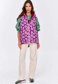 Kobieta ubrana w fioletową kwiecistą kamizelkę pikowaną na zielone długie rękawy, beżowe spodnie oraz zielono-fioletowe trampki, stojąca na tle białym.