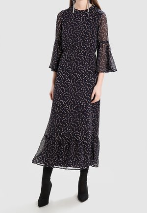 Femme portant une longue robe noire avec de petits motifs floraux blancs et rouges, des manches cloche transparentes et des bottines noires sur un fond clair.