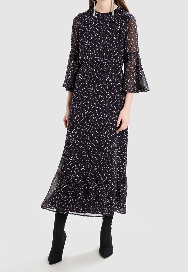 Femme portant une longue robe noire avec de petits motifs floraux blancs et rouges, des manches cloche transparentes et des bottines noires sur un fond clair.