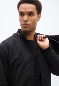 Jeune homme aux cheveux courts et bouclés, portant une veste noire par-dessus une chemise noire, tenant le col de la veste et regardant sur le côté.