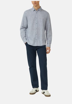 Mężczyzna w jasnoszarej, paskowanej koszuli z długim rękawem i guzikami, ciemnoniebieskich spodniach i białych trampkach z czarnymi akcentami, stoi z jedną ręką w kieszeni.