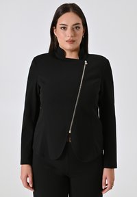 Zwarte jas met lange mouwen, een hoge kraag en een asymmetrische voorkant. Heeft een gouden rits aan de zijkant en een gladde stoftextuur.