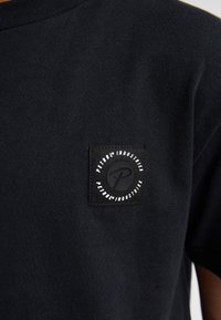 Zwart katoenen T-shirt met een vierkant zwart label met witte tekst en logo. Korte mouwen en een ronde hals. Zachte textuur.