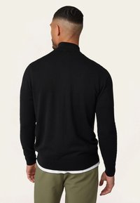 Zwarte gebreide trui met een hoge kraag, aansluitende lange mouwen en geribbelde manchetten. Draagt over een witte shirt, gecombineerd met olijfgroene broek.