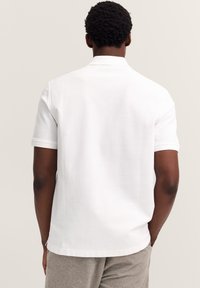 Mann mit dunklem, lockigem Haar, der ein schlichtes weißes Polohemd mit kurzen Ärmeln und hellgraue Hosen trägt, mit den Händen in den Taschen stehend und von hinten blickend.