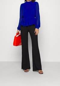Blusa blu a maniche lunghe con vita arricciata, abbinata a pantaloni neri a gamba larga. Una borsa lucida rossa completa l'outfit.