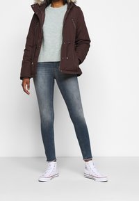 Vinröd isolerad jacka med fuskpäls, ljusgrön tröja, blåa tajta jeans och vita höga sneakers med röda detaljer.