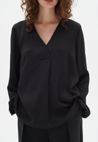 Blusa nera con scollo a V, realizzata in tessuto morbido, con maniche lunghe e una vestibilità comoda. Dettaglio di drappeggio discreto sul davanti.