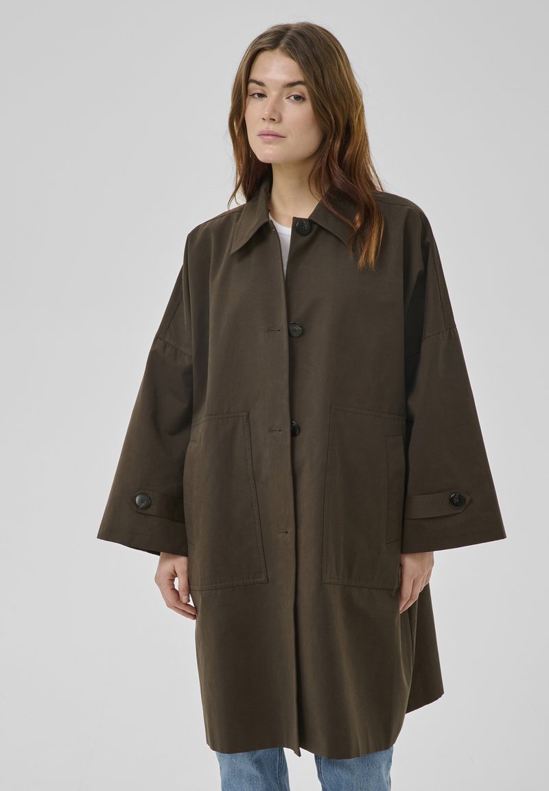 Jonge vrouw die een oversized donkerbruine jas met grote knopen en ruime voorzakken draagt, en een blauwe spijkerbroek, staand tegen een effen lichte achtergrond.