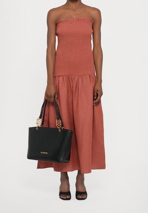 Kvinde iført en skulderfri, rustrød midikjole, der holder en sort Love Moschino shopper med gulddetaljer, iført sorte åbne høje hæle.