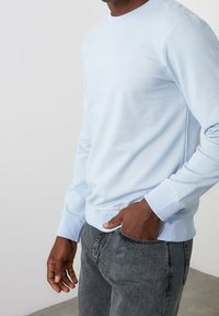 Pull à col rond bleu clair en tissu lisse, associé à un jean gris. Le pull présente des poignets et un bas côtelés pour une coupe ajustée.