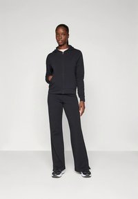 Črna kapucar z zadrgo, z gladko teksturo in žepom kenguru, kombiniran s črnimi razširjenimi hlačami. Superge imajo belo-črni podplat.