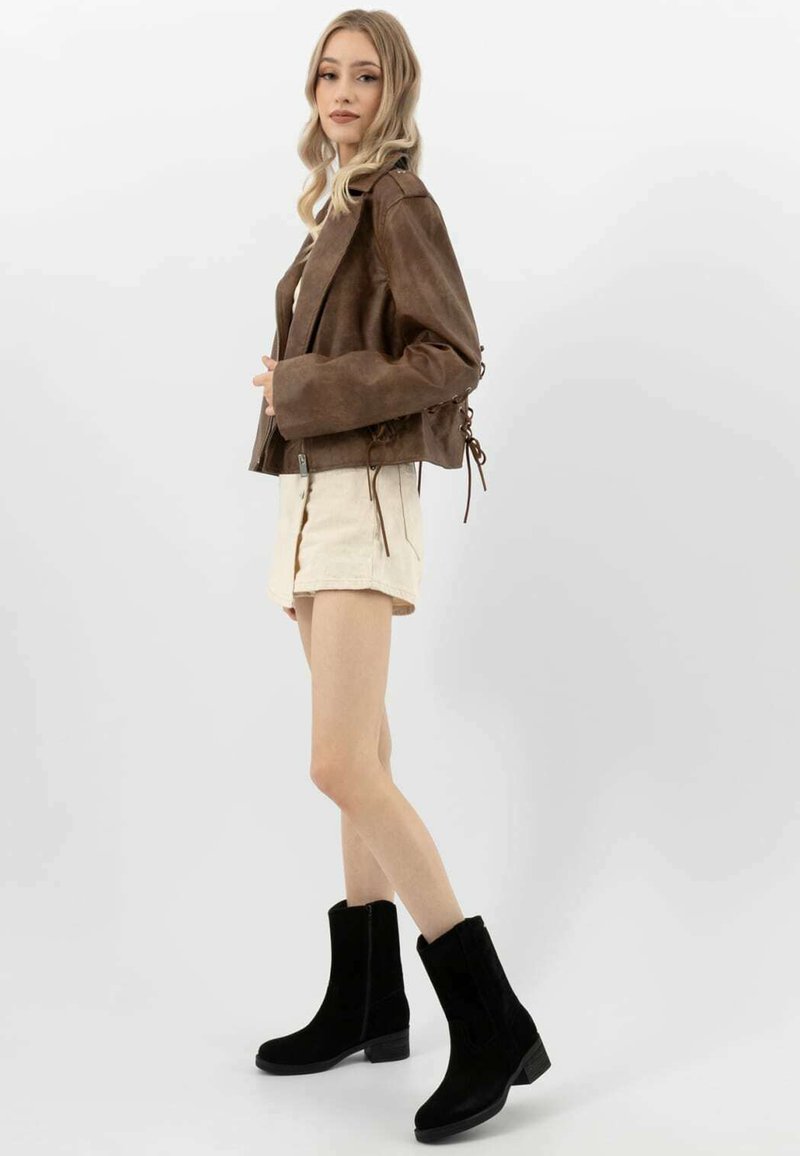 Donna in piedi di lato che indossa una giacca in suede marrone, una gonna beige e stivali neri fino al polpaccio su uno sfondo bianco uniforme.