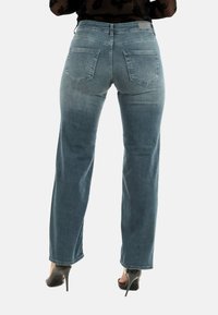 Donna che indossa jeans sbiaditi a gamba dritta blu e tacchi alti neri, vista da dietro su uno sfondo neutro.
