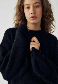 Pulover negru, pufos, din tricot, cu guler înalt și mâneci largi, supradimensionate. Țesătura pare moale și texturată. Fără modele sau accente vizibile.