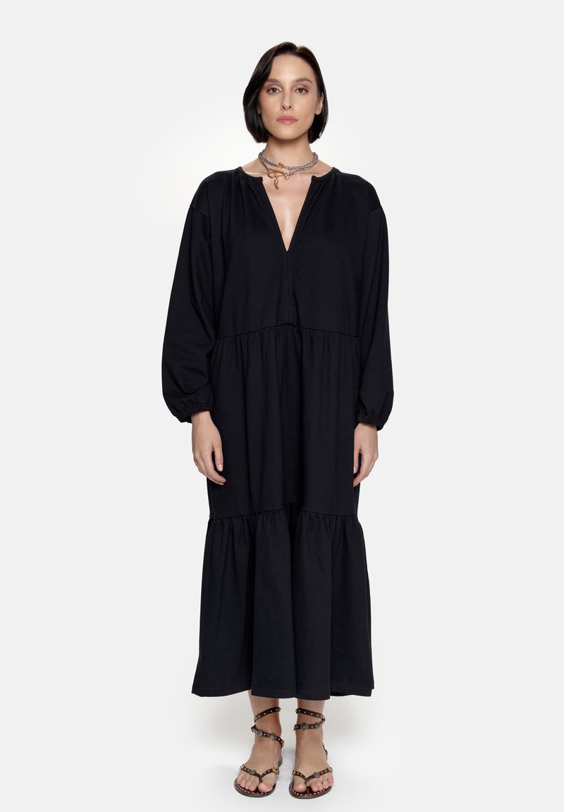 Czarna sukienka z długimi rękawami, wykonana z miękkiego materiału, z dekoltem w kształcie litery V oraz warstwową spódnicą o luźnym kroju. W zestawie z sandałami na paski i biżuterią.