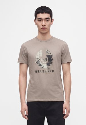 Ung man som bär en beige kortärmad t-shirt med en blekt svart grafik och texten "BELSTAFF", stående mot en enfärgad vit bakgrund.