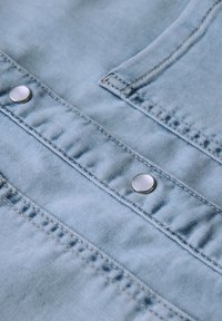Tessuto in denim blu chiaro con una superficie liscia, caratterizzato da bottoni in metallo argentato e cuciture visibili lungo i bordi e le cuciture.