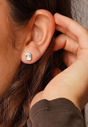 Silver stud earrings with an emerald-cut clear stone, featuring a simple design and smooth surface, worn in a pierced ear.