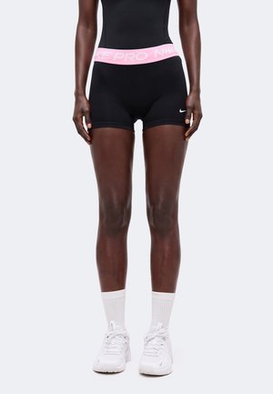 Fekete Nike Pro rövidnadrágot viselő személy, rózsaszín derékpánttal, fehér felnőtt zoknival és fehér sportcipővel, egyszínű háttér előtt áll.