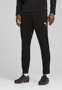 Calças de desporto pretas com detalhes laranja nas laterais, apresentando um ajuste solto e cintura elástica, combinadas com botas de futebol pretas com detalhes em laranja.