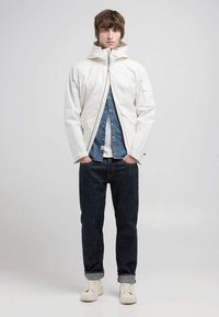 Chaqueta ligera blanca con cuello alto, cremallera frontal y bolsillos laterales, llevada sobre una camisa de mezclilla y vaqueros oscuros, acompañada de zapatillas blancas.