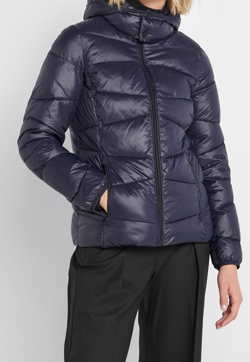 Donna che indossa una giacca a vento trapuntata blu navy con zip frontale e pantaloni neri, mano nella tasca della giacca, su uno sfondo neutro.